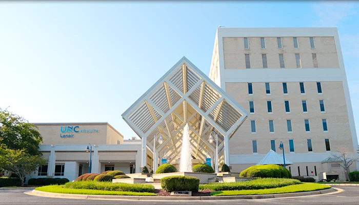 Modern building with a geometric entrance structure, surrounded by manicured bushes and a fountain. Clear sky enhances the welcoming ambiance.