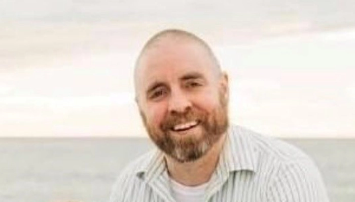 A warm, professional portrait of professional. He has short hair, is wearing a collared shirt and is smiling directly at the camera against a bright outdoor backdrop