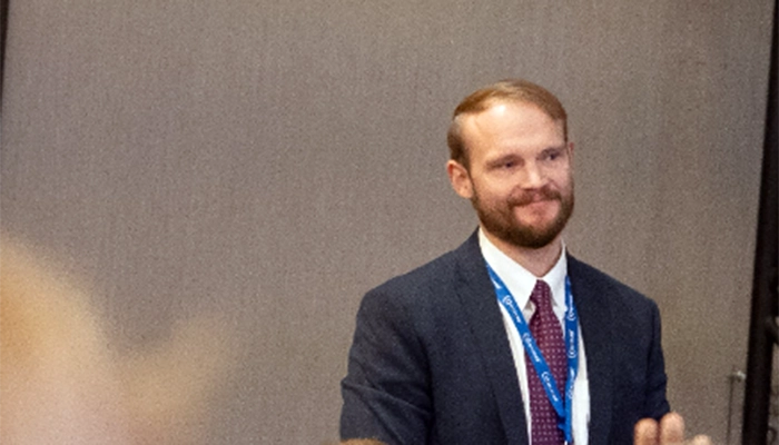 A man with a beard, wearing a suit and tie, stands smiling with a blue lanyard around his neck against a plain background, conveying a professional and friendly demeanor.