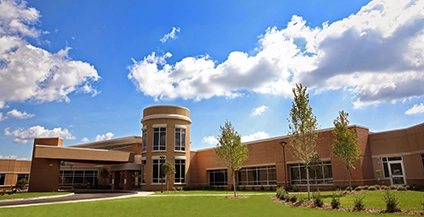 Brick building with large windows and a cylindrical entrance under a vibrant blue sky with scattered clouds. Trees and a grassy area in front.