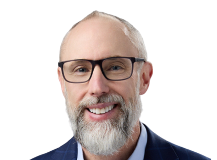 Bobby Ryan, with glasses and a white beard, wearing a blue suit and a light blue shirt, against a plain white background. The mood is professional and approachable.