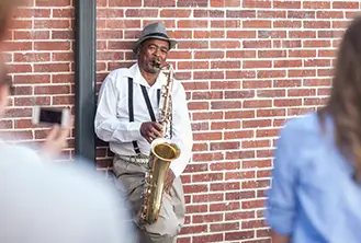 man playing saxophone in the street, leaning on a brick wall