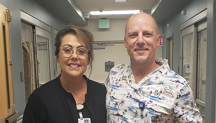 Two people stand in a hospital hallway, smiling. One wears a black uniform and the other a tropical-patterned shirt, conveying a friendly atmosphere.