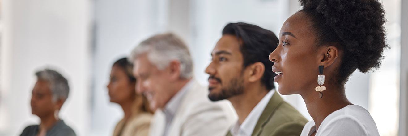 A diverse group of five people in a row, attentively looking forward. The focus is on a woman in the foreground, expressing interest and engagement.