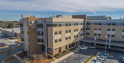 Aerial view of a modern, multi-story medical center with a brown and beige facade. Cars parked outside. Clear sky, conveying a professional, calm atmosphere.