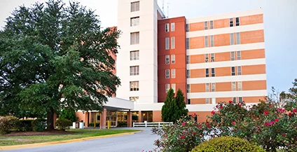 Exterior of Onslow Memorial Hospital, a tall white and tan building with orange accents, surrounded by green trees and flowering bushes.