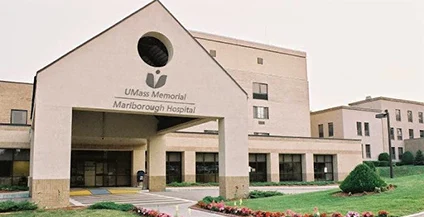 Front view of UMass Memorial Marlborough Hospital, featuring a beige and brown building with a covered entrance. It is surrounded by green lawns and bushes.