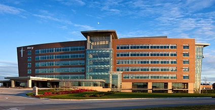 Modern brick and glass building under a clear blue sky, featuring a covered walkway, surrounded by landscaped greenery and an empty paved driveway.