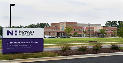 Sign for Novant Health Clemmons Medical Center in foreground, with a red-brick building and multiple flags in the background under a cloudy sky.