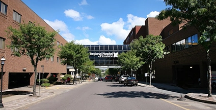 Street view of the Regional Hospital of Scranton, with a skywalk connecting buildings. Trees line the street under a clear blue sky with scattered clouds.