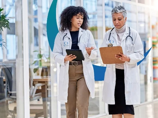 Two doctors in white coats walk in a corridor, discussing notes on a clipboard and tablet. The setting is bright and professional.