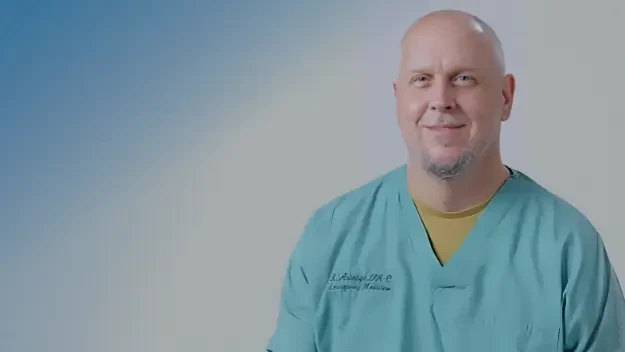 A man in a blue medical scrub smiles softly, conveying a calm and approachable demeanor. The background is a gradient of blue to white.