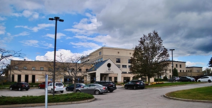 A large, contemporary hospital building under a partly cloudy sky, with several parked cars in the front lot and trees surrounding the entrance.