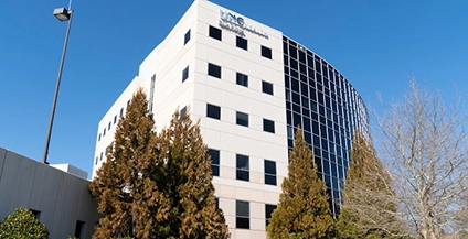 Modern office building with a mix of white paneling and reflective glass windows, surrounded by evergreen trees, under a clear blue sky.