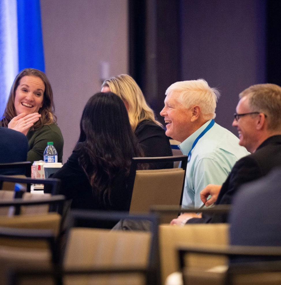 A group of people sit at a conference table, smiling and conversing. The room has a warm and friendly atmosphere with natural lighting.