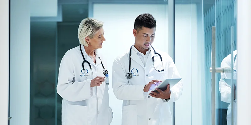 Two doctors in white coats walk down a hospital corridor, discussing notes on a tablet. Their expressions are focused and professional.