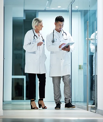 Two doctors in white coats walk down a hospital corridor, discussing notes on a tablet. Their expressions are focused and professional.