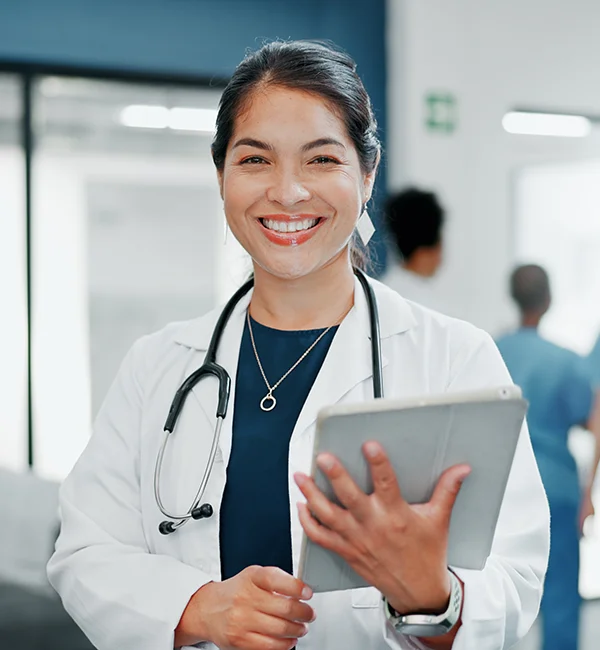 Smiling female doctor with a stethoscope and tablet, wearing a white coat. Background shows a busy hospital setting with medical staff in scrubs.