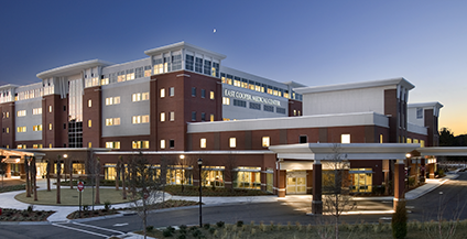 Modern hospital building at dusk with illuminated windows, red-brick facade, and multiple stories. The sky is a soft blue-gray, creating a calm atmosphere.