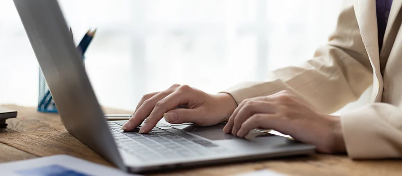 Close-up of a person’s hands typing on a laptop keyboard at a wooden desk in a bright, professional setting