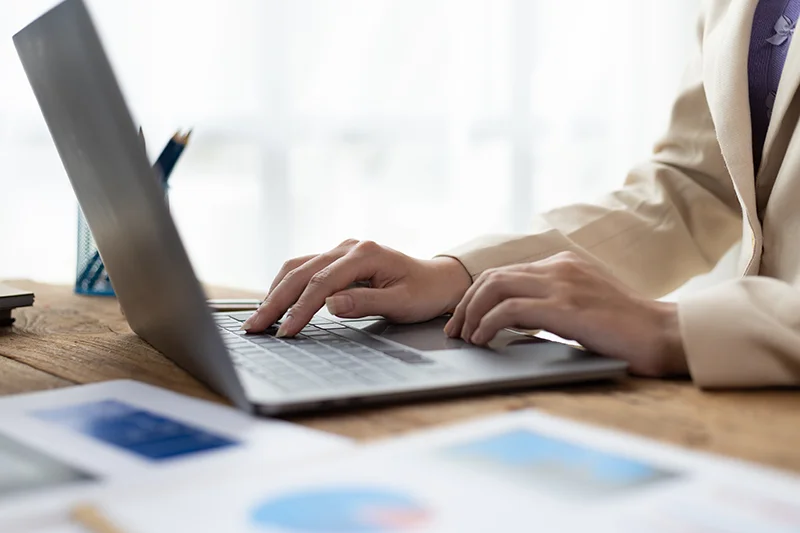 Close-up of a person’s hands typing on a laptop keyboard at a wooden desk in a bright, professional setting