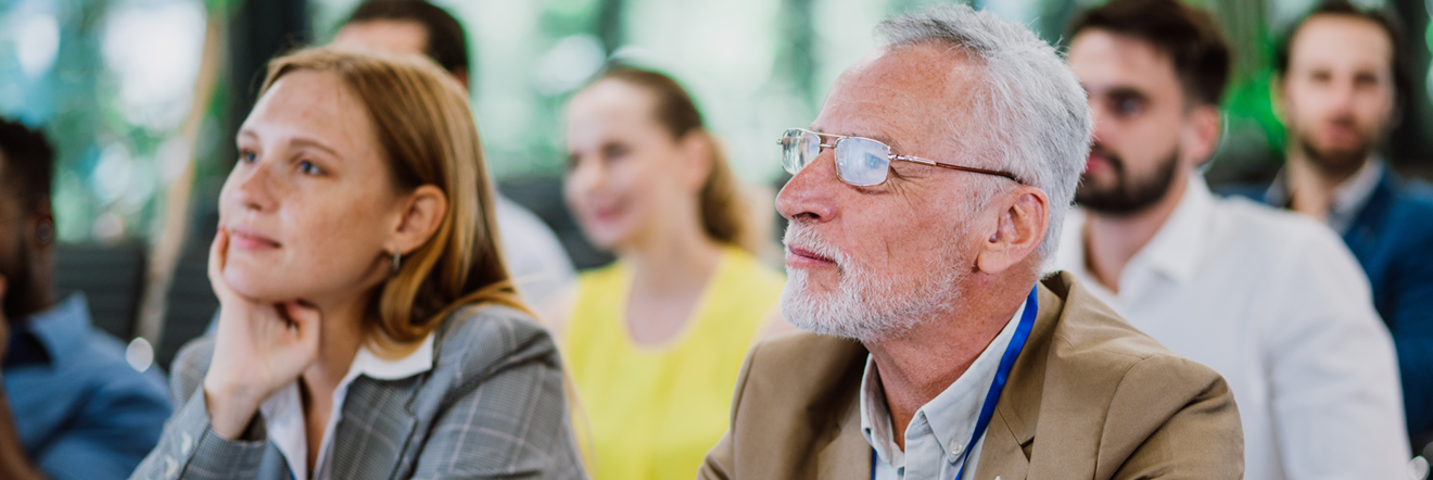 A diverse group of people attentively listening in a seminar. A woman in a gray suit and an older man with glasses appear focused and engaged.