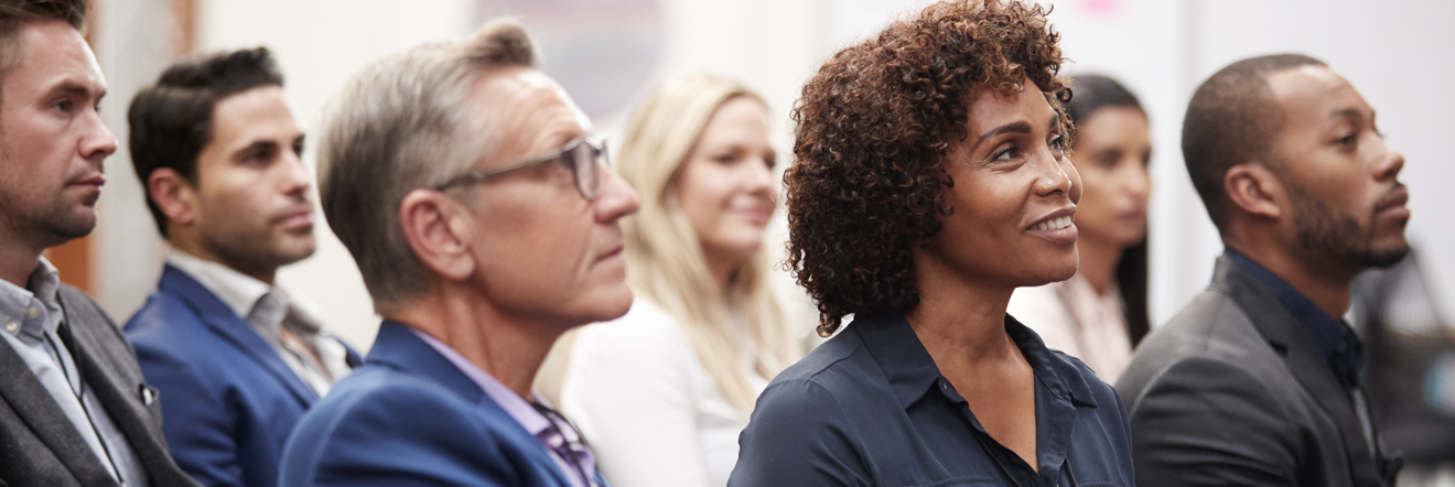 Audience seated in a conference setting, focused intently. A woman in the foreground smiles. The mood is attentive and engaged.