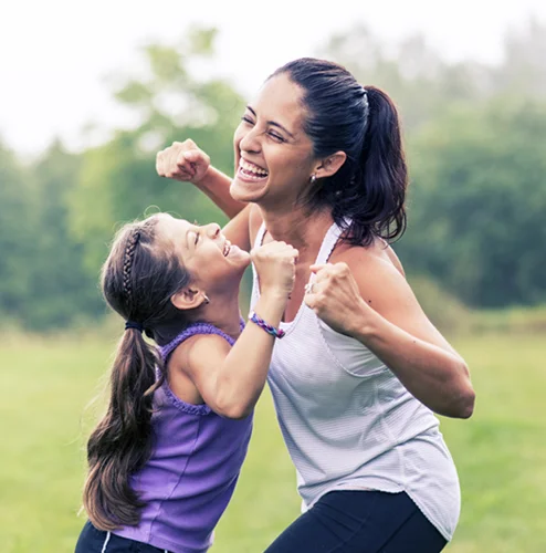A joyful mother and daughter in athletic wear celebrate outdoors in a grassy park. Both smile brightly, fists raised, conveying excitement and happiness.