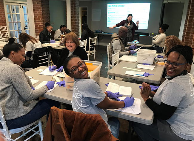 Group of people in a workshop setting, seated at tables with notebooks. They are wearing purple gloves. A woman stands presenting. Smiling, collaborative atmosphere.