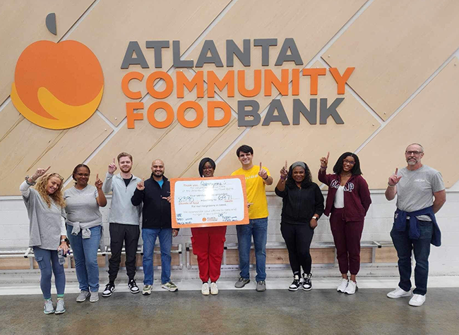 A diverse group of ten people stands smiling in front of the Atlanta Community Food Bank logo, holding a large check. They appear cheerful and supportive.