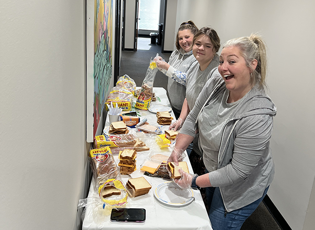 Three women in gray shirts happily make sandwiches on a long table in a hallway. Bread, cheese, and meat are spread out. A phone lies nearby.