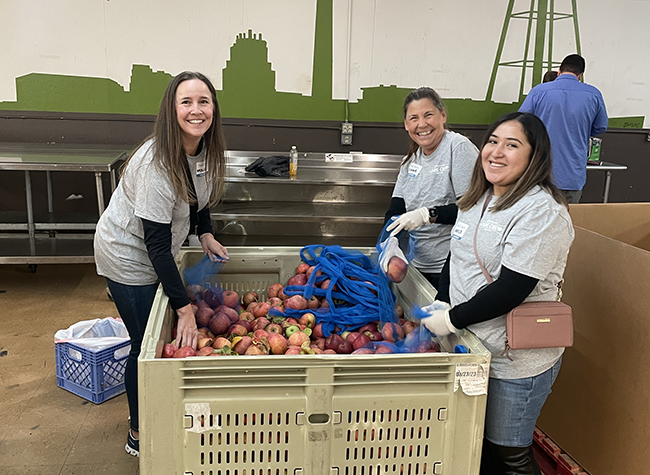 Three women in gray shirts smile while sorting apples in a large crate at a food bank. The mood is cheerful, conveying teamwork and community service.