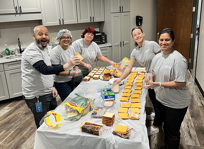 Five people in matching gray shirts joyfully make sandwiches on a long table covered with bread, cheese, and deli meats in a kitchen setting.