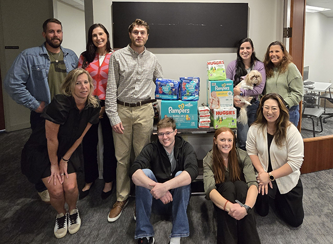 A group of ten people smiling and posing indoors with stacked packages of Pampers and Huggies diapers. One person holds a small dog, conveying a cheerful and collaborative atmosphere.