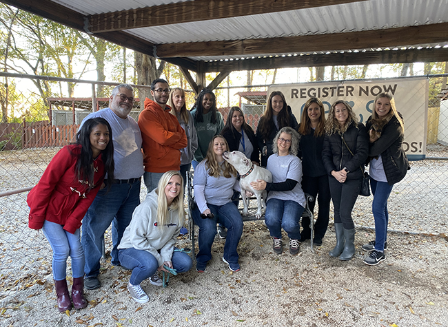 A diverse group of 14 people and a dog pose happily in a fenced outdoor area under a roof. They stand and kneel, smiling, with a banner behind them.