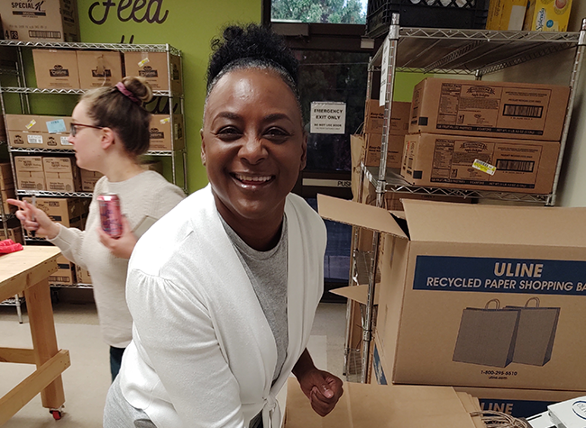 Smiling woman in a food bank stands beside stacked boxes, wearing a white sweater. Another person in the background holds a soda can. The scene is lively and joyful.