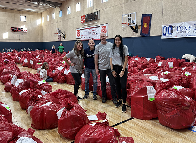 A group of four people stands on a gym floor surrounded by large red bags. The scene is busy, with numerous bags filling the area. The mood is positive and collaborative.