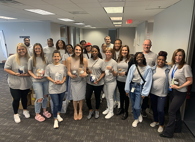 A diverse group of 18 people smiling indoors, casually dressed, holding small gifts. The setting is an office hallway, conveying a cheerful, team-building atmosphere.