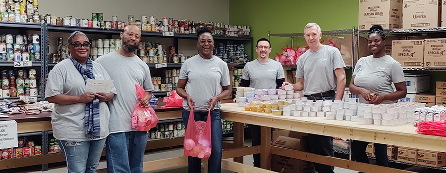 A group of six smiling individuals in gray shirts volunteering at a food bank, organizing canned goods and packing items in red bags. Shelves are stocked behind them.