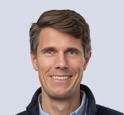A bright, approachable professional portrait of a man. He has a wide, warm smile and brown hair. He is looking directly at the camera against a neutral grey studio backdrop.