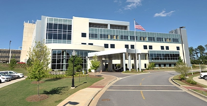 Modern hospital building with large windows, a curved entrance, and a flagpole with a waving American flag. Landscaping includes greenery and trees.