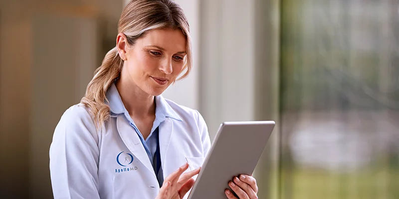 A female healthcare professional in a white lab coat with an ApolloMD logo, smiling while looking at and interacting with a silver tablet in a bright, modern clinical setting.