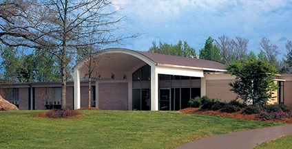 Modern building with an arched roof, large glass windows, and light brick walls. Set against a blue sky with surrounding trees and lush grass.