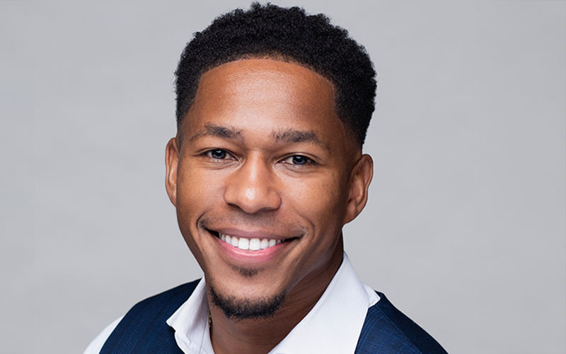 Professional headshot of Dany Accilien, a man wearing a white medical lab coat over a dark shirt and tie, set against a neutral studio background.