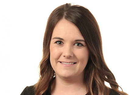 A warm, professional portrait of professional. She has wavy brown hair, is wearing a blazer, and is smiling directly at the camera against a bright backdrop