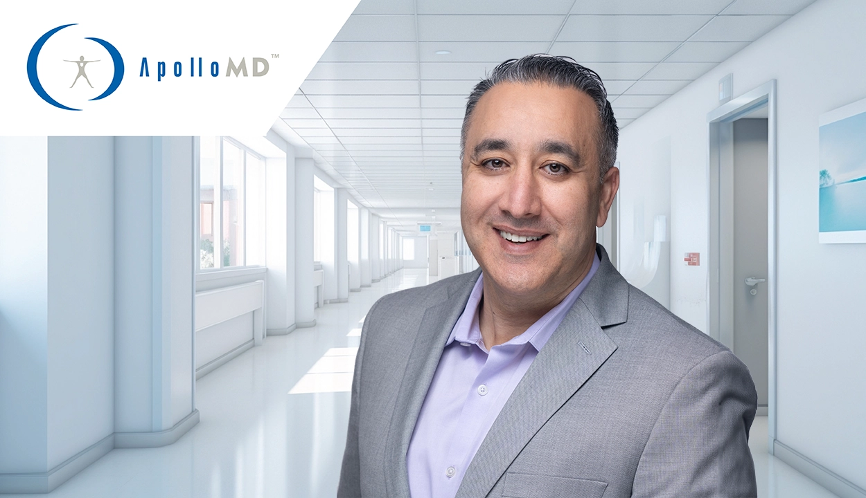 A professional portrait of a man in a dark suit and blue tie, standing in a brightly lit, modern medical office hallway with glass walls and contemporary lighting.