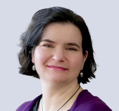 A professional headshot of a woman with dark hair, wearing a white blouse and a dark blazer, set against a blurred indoor office background.