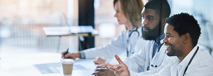 Three doctors in white coats sit at a conference table, engaged in discussion. The setting is bright and professional, conveying collaboration.