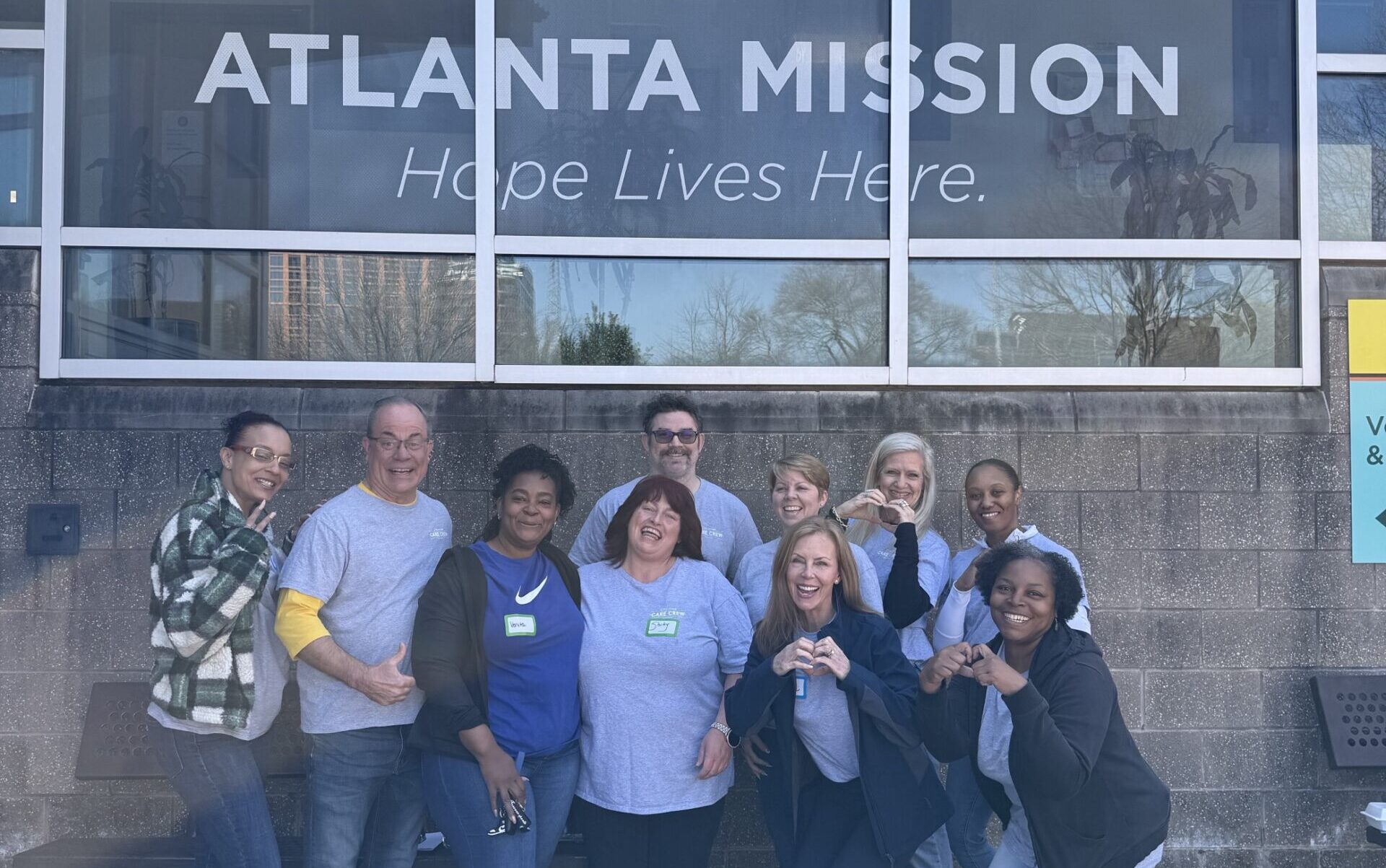 A group of smiling people pose together outside the Atlanta Mission building. They appear cheerful, with some making heart symbols with their hands.