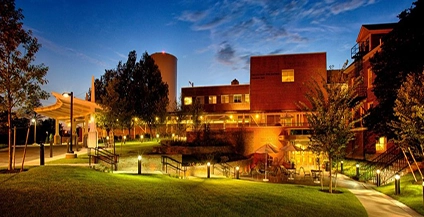 Modern brick building and surrounding lush lawns illuminated by warm lights at dusk. Trees and a deep blue sky add a serene ambiance.